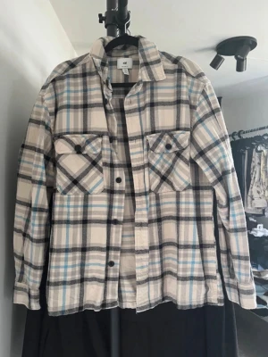 H&M Relaxed Fit overshirt/flanellskjorta - H&M Relaxed Fit overshirt i mjuk bomullsflanell med beige, grått, svart och ljusblått rutmönster. Två bröstfickor med lock, klassisk skjortkrage och en avslappnad passform. Perfekt som lager-på-lager under höst och vår eller öppen över en T-shirt. Fint skick.