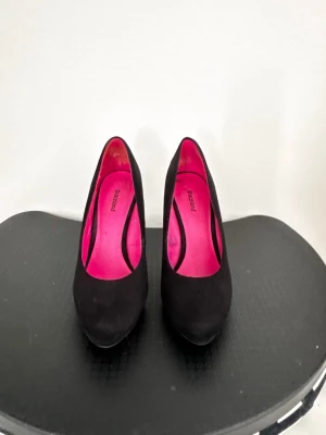 Svarta klassiska pumps med platå - Säljer ett par svarta pumps med hög klack och lätt platå framtill. Skorna har en stilren design med rundad tå och insida i starkt rosa färg. Materialet ser ut att vara mockaimitation som ger en mjuk känsla. Perfekta för dig som vill ha en tidlös och elegant look.