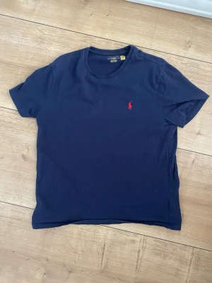Mörkblå t-shirt från Polo Ralph Lauren - Klassisk mörkblå t-shirt från Polo Ralph Lauren med den ikoniska röda logotypen broderad på bröstet. T-shirten har rund halsringning och korta ärmar. Tillverkad i mjuk bomull för en skön känsla och enkel stil.