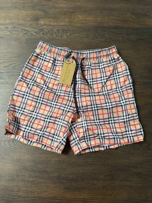 Rutig shorts från Burberry med snörning - Snygga shorts från Burberry i klassiskt rutigt mönster med beige, svart, vitt och röda detaljer. Shortsen har elastisk midja och svart snörning framtill. Perfekta för sommaren och ger en trendig look.