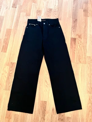 Svarta raka jeans med hög midja - Snygga svarta jeans i storlek W27/L30 med  rak passform och hög midja. Klassisk femficksmodell med silverfärgade nitar och knapp. Jeansen har breda ben och är tillverkade i ett kraftigare denimtyg. Perfekta för en clean och stilren look.