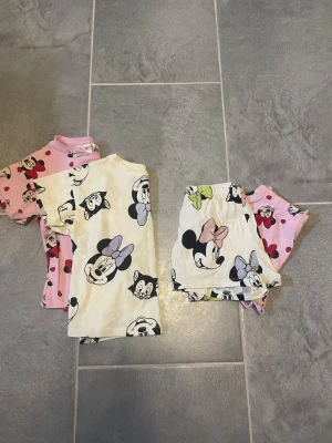 Två pyjamas-set med Minnie Mouse - Säljer två söta pyjamas-set för barn med Minnie Mouse-tryck. Ena setet är rosa med röda hjärtan och Minnie-motiv, det andra är vitt med färgglada detaljer och Minnie-tryck. Båda seten består av t-shirt och shorts i mjuk bomull, perfekta för mysiga kvällar.