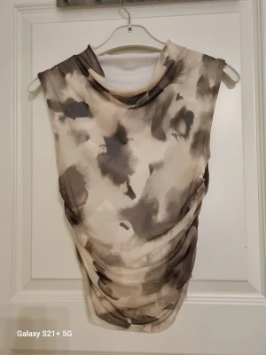Mönstrad ärmlös topp från Kappahl - Säljer en ärmlös topp från Kappahl i beige och grå toner med abstrakt mönster. Toppen har draperad halsringning och lätt rynkade sidor för en snygg siluett. Perfekt att styla med jeans eller kjol för en trendig look.
