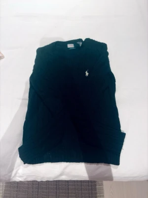 Mörkblå kabelstickad tröja Polo Ralph Lauren - Kabelstickad tröja i mörkblått från Polo Ralph Lauren med klassisk vit broderad logga på bröstet. Tröjan har rund halsringning och ribbade muddar vid ärmslut och nederkant. Perfekt för en stilren och tidlös look.