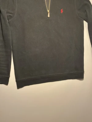 Svart half-zip tröja från Polo Ralph Lauren - Klassisk svart tröja med half-zip från Polo Ralph Lauren. Tröjan har en hög krage, dragkedja med läderdetalj och den ikoniska röda logon broderad på bröstet. (Säljs pågrund av att den sällan används mer) ”Vid minsta frågor eller funderingar så är det bara att höra av sig” 😁
