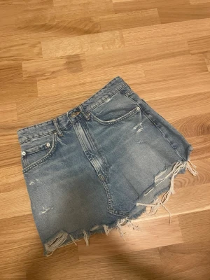 Blå jeanskjol med fransig kant från Zara - Säljer en blå jeanskjol från Zara i storlek S. Kjolen är kort med rå, fransig kant och har klassiska fem fickor. Perfekt för dig som gillar en avslappnad och trendig stil.