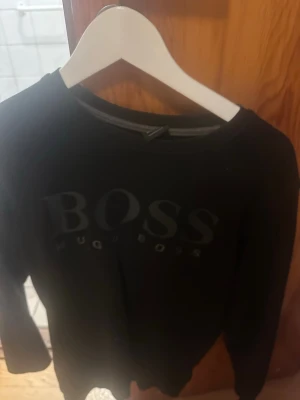 Svart sweatshirt från Hugo Boss - Snygg svart sweatshirt från Hugo Boss med stor logga framtill. Tröjan har rund halsringning och långa ärmar. Perfekt för en stilren och avslappnad look.