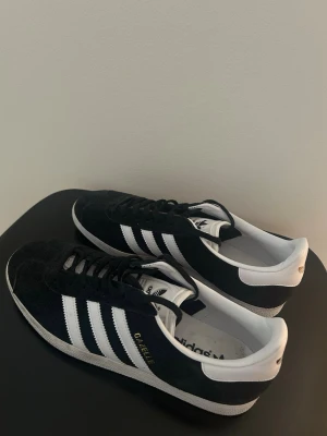 Adidas Gazelle svart/vit mocka - Jag säljer mina adidas Gazelle sneakers i svart mocka med vita detaljer och de klassiska tre ränderna på sidan. Låg modell med vit sula och vit hälkappa. Priset är inte spikat! Använda 2 gånger pga fel storlek. Nypris:1200