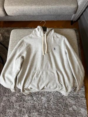 Ljusgrå hoodie från Polo Ralph Lauren - Snygg ljusgrå hoodie från Polo Ralph Lauren med klassisk huva och vita dragsnören. Har en känguruficka framtill och den ikoniska lilla loggan broderad på bröstet. Perfekt för en chill och avslappnad stil.