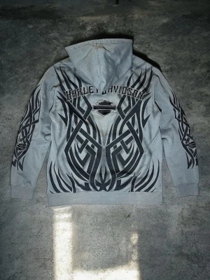 Grymt Skick Äkta Vintage Harley Davidson Tribal All Over Print Hoodie Stl M - Äkta vintage Harley Davidson huvtröja i absolut toppskick. Extremt sällsynt modell med all over tribalmönster över rygg och ärmar samt broderad Bar and Shield logga. Perfekt för Y2K grunge biker och cyberestetik. Märke: Harley Davidson, Storlek: M, Färg: Tvättad grå och svart, Material: 80% bomull 20% polyester, Skick: Helt grymt vintageskick utan hål fläckar eller slitage på tryck. Djur och rökfritt hem. Skickas snabbt och spårbart.
