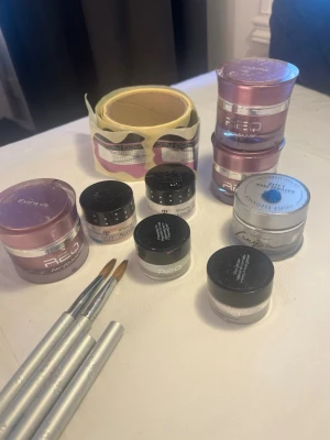 Nagelkit med akrylpulver och penslar - Säljer ett komplett nagelkit med flera burkar akrylpulver i olika storlekar och färger, bland annat vitt och rosa. Inkluderar två silverfärgade nagelpenslar och nagelformar på rulle. Perfekt för dig som vill skapa egna akrylnaglar hemma.
