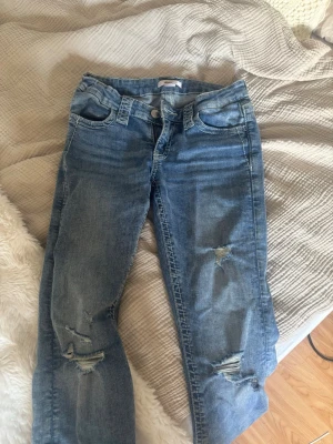 Blå slitna jeans med broderad ficka - Säljer ett par ljusblå jeans med slitningar på benen och snyggt broderad bakficka med paljettdetaljer. Jeansen har klassisk femficksmodell och raka ben. Perfekta för dig som gillar en avslappnad och trendig stil.