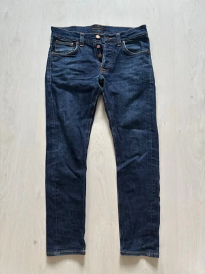Mörkblå jeans från Nudie Jeans - Snygga mörkblå jeans från Nudie Jeans med raka ben och klassisk femficksmodell. Jeansen har orange kontrastsömmar och knäppning med kopparfärgade knappar. Tillverkade i slitstark denim, perfekta för en avslappnad och trendig look.