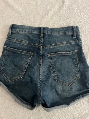 Blå denimshorts med upprullad kant - Snygga blå jeansshorts i klassisk modell med upprullad kant och femficksdesign. Shortsen har bälteshällor, hög midja, knapp och dragkedja framtill samt två bakfickor. 