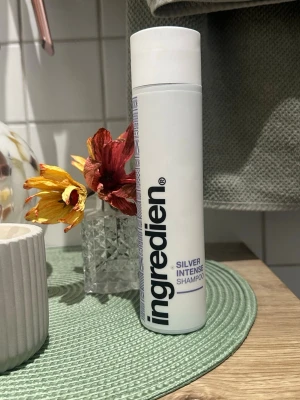 Ingredien Silver Intense Shampoo - Ungefär hälften kvar. Ny.pris 379kr Silver Intense Shampoo från Ingredien i en vit, cylinderformad plastflaska med svart och lila text. Perfekt för att neutralisera gula toner i blont eller grått hår. Flaskan har ett stilrent och modernt utseende.