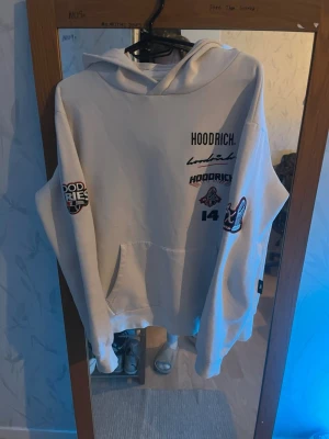 Vit hoodie från Hoodrich med tryck - Säljer en vit hoodie från Hoodrich med flera coola tryck och broderade detaljer på både fram- och baksida samt ärmar. Hoodien har stor huva och känguruficka framtill. Perfekt för dig som gillar streetwear och vill sticka ut.