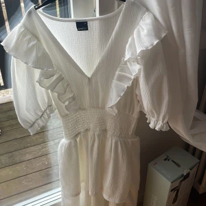 Vit volangklänning från Gina Tricot - Supersöt vit kortklänning från Gina Tricot med v-ringning, volanger på axlarna och puffärmar. Klänningen har smock i midjan och är tillverkad i ett lätt, krinklat bomullstyg som ger en luftig känsla. Perfekt för sommarens alla tillfällen! Använt endast en gång och klänningen är som ny! 