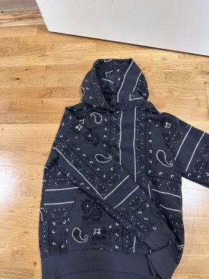 Svart hoodie med bandanamönster Zara - Säljer en svart hoodie från Zara i storlek M med vitt bandanamönster och detaljer som stjärnor och paisley. Hoodien har huva och ribbade muddar vid ärmslut och nederkant. Perfekt för dig som gillar streetwear och vill sticka ut lite extra.