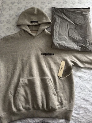Essentials hoodie Fear of God - Snygg grå hoodie från Essentials Fear of God med svart logga på bröstet. Aldrig använd, dustbag och tagsen kommer med. I storlek S men sitter oversized så passar även M. Klassisk känguruficka framtill och stor huva. Tillverkad i mjukt bomullsmaterial som känns riktigt skönt mot huden. Perfekt för en avslappnad och trendig streetwear-look.