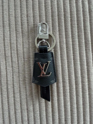  Vuitton nyckelring i svart läder - Snygg nyckelring från  Vuitton i svart läder med LV-logga i metall framtill. Har både ring och karbinhake i silverfärgad metall, perfekt att fästa på väskan eller nycklarna. Klassisk och stilren accessoar med exklusiv känsla.