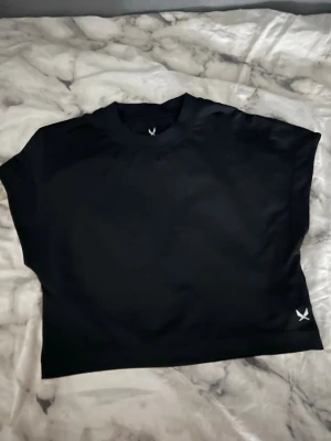 Svart croppad topp med tryck - Snygg svart croppad topp med rund hals och korta ärmar. Toppen har ett diskret vitt tryck nertill på ena sidan. Perfekt till jeans eller kjol för en trendig look.