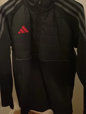 Svart träningsjacka från Adidas - Säljer en svart träningsjacka från Adidas med halv dragkedja och röda Adidas-loggan på bröstet. Jackan har gråa ränder på axlarna och quiltad struktur framtill. Perfekt för träning eller uppvärmning.