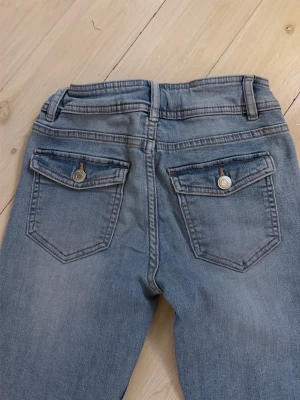 Ljusblå bootcut jeans med ficklock från kapphal - Säljer ett par ljusblå jeans bootcut och två bakfickor med lock och knapp. Jeansen har klassisk femficksdesign, bälteshällor och är gjorda i denim. Perfekta för en avslappnad och trendig look. Har lite pytte lite lim i en av fickorna som är hårt men man ser inte det är mycket ljusare i verkligheten vet storlek 152 cm fast jag skrev bara 30 där nere för behövde välja nån