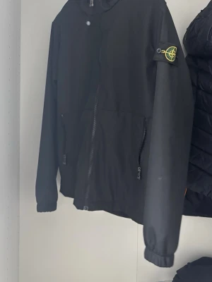 Svart vindjacka från Stone Island - Snygg svart vindjacka från Stone Island med klassisk patch på ärmen. Jackan har dragkedja framtill, två sidofickor med dragkedja och ribbade muddar vid ärmslut. Perfekt för dig som vill ha en stilren och ikonisk look.