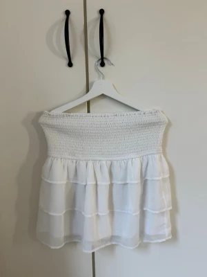 Vit volangkjol med smock - Söt vit kortkjol med bred smock i midjan och flera lager volanger. Kjolen har en luftig och lätt känsla, perfekt för varma dagar. Materialet känns som bomull eller polyester och ger en flowy look.