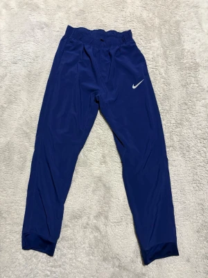 Blå Nike Dri-FIT träningsbyxor - Säljer ett par blå Nike Dri-FIT träningsbyxor med resår i midjan och justerbar dragsko. Byxorna har en smal passform och är tillverkade i ett lätt, snabbtorkande material som passar perfekt till träning eller sportaktiviteter.