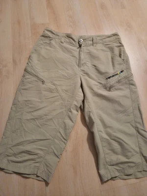 Beige shorts från Tenson - Säljer ett par beige shorts från Tenson storlek S med dragkedjeförsedda fickor på sidorna och broderad logga. De har normal passform, bälteshällor och är tillverkade i ett lätt syntetmaterial som passar perfekt för varma dagar.