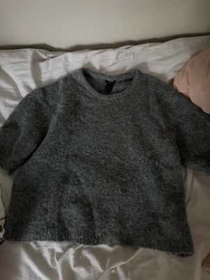 Grå fluffig stickad tröja - Mysig grå stickad tröja med rund hals och kortare ärmar. Tröjan har en fluffig och mjuk textur, perfekt för lager-på-lager och chill dagar. Enkel design som passar till det mesta och ger en avslappnad vibe.