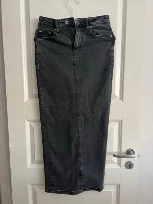 Grå lång jeanskjol med slits - Snygg lång jeanskjol i mörkgrå denim med klassiska fickor och bälteshällor. Kjolen har en rak passform och en slits framtill som ger extra rörelsefrihet. Perfekt att styla med sneakers eller boots för en cool look.