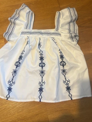 Vit topp med blå brodyr från SHEIN - Supersöt vit topp från SHEIN med blå broderade blommor och vågiga detaljer på axelbanden. Är perfekt för sommaren och varma väder. Broderierna ger en boho-vibe och toppen har en luftig passform.
