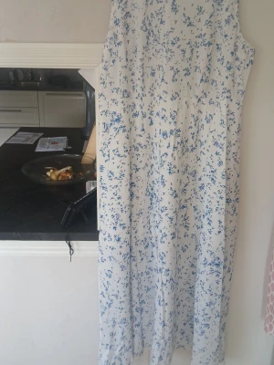 Vit långklänning med blått blommönster - Vit långklänning med blått blommigt mönster. Klänningen är ärmlös och har en luftig, rak passform som passar perfekt till varma dagar. Tillverkad i ett lätt material som känns svalt mot huden.