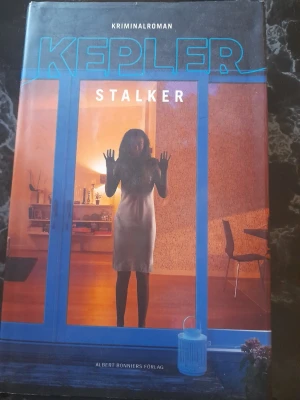 Stalker av Lars Kepler - En spännande kriminalroman av Lars Kepler med ett omslag som visar en kvinna i motljus genom ett fönster. Boken är utgiven av Albert Bonniers Förlag och har en mörk, mystisk design med blå och orange toner. Perfekt för dig som gillar nervkittlande thrillers.