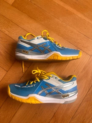 Blå och gula Asics inomhusskor - Säljer ett par sportiga Asics inomhusskor i blått, gult, svart och vitt. Skorna har gula snören, slitstark gummisula och ventilerande meshmaterial. Perfekta för handboll, volleyboll eller andra inomhussporter. Snygg design med Asics-logga på sidan.