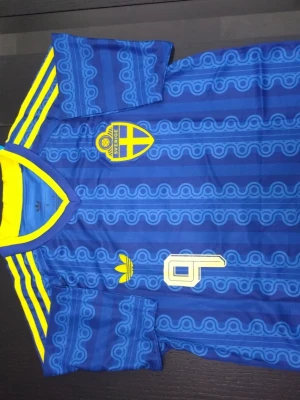 Blå Sverige fotbollströja Adidas - Retroinspirerad blå fotbollströja från Adidas med Sveriges landslagsemblem på bröstet och gula detaljer. Tröjan har gula ränder på axlarna, mönstrad design och nummer 9 på framsidan. Perfekt för dig som vill sticka ut på match eller träning.