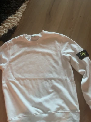 Vit sweatshirt från Stone Island - Säljer en clean vit sweatshirt från Stone Island med den klassiska svarta patchen med gul logga på ärmen. Tröjan har rund halsringning och ribbade muddar. Perfekt för dig som gillar streetwear och vill ha en stilren look.