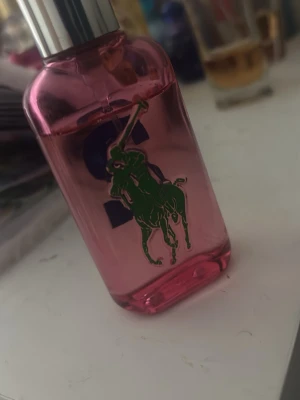 Rosa parfymflaska från Ralph Lauren - Säljer en snygg parfymflaska från Ralph Lauren i genomskinligt rosa glas med silverfärgat lock. Flaskan har en grön logga med en polospelare och stora blå siffror på framsidan. Perfekt för dig som gillar sportig och fräsch design.