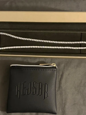 Tennishalsband Kejsar  - Tennishalsband från kejsar. Den har aldrig använts och var en present som tyvärr inte är i min smak. Den kommer i nytt skick utan orginalförpackning, men levereras med smyckefodralet med dragkedja i bilden (den med kejsar loggan). 