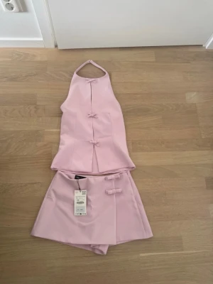 Ljusrosa set med topp och kjolshorts - Säljer ett ljusrosa set med halterneck-topp och matchande kjolshorts. Toppen har dekorativa knytningar framtill och kjolshortsen har en liknande detalj i midjan. Materialet ser ut att vara slätt och lätt, perfekt för en trendig look.