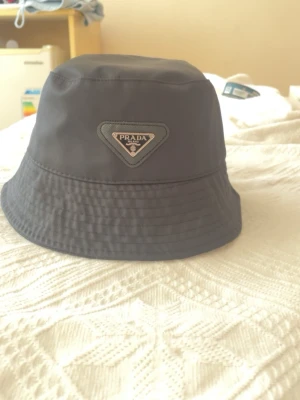 Svart bucket hat från Prada - Snygg svart bucket hat från Prada med det klassiska trekantiga metallogot på framsidan. Hatten är tillverkad i ett glansigt, vattenavvisande material som ger en modern och clean look. Perfekt accessoar för att lyfta din outfit.