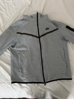 Grå Nike hoodie med dragkedja - Snygg grå hoodie från Nike med hel dragkedja och svart logga på bröstet. Hoodien har svarta detaljer längs bröstet och ärmarna samt fickor på sidorna. Tillverkad i mjukt sweatshirtmaterial och har en huva som går att dra upp för extra stilpoäng.