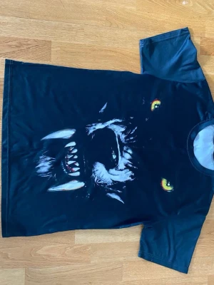 Svart t-shirt med vargtryck - Cool svart t-shirt med stort tryck av en vargs ansikte på framsidan. T-shirten har korta ärmar och rund hals. Trycket har detaljer i vitt, gult och rött som gör motivet riktigt iögonfallande. Perfekt för dig som gillar djurmotiv och streetwear.