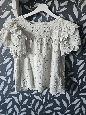 Grön blus med broderade volangärmar - Söt vit blus från Monki med broderade blommönster och volangprydda korta ärmar. Blusen har rund halsringning och är tillverkad i ett lätt, luftigt material med hålbroderier som ger en romantisk känsla.