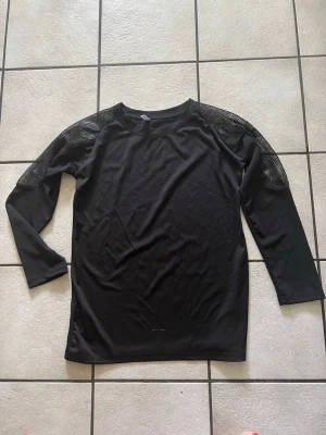 Svart mesh-detaljerad blus storlek M/L - Snygg svart blus med lång ärm och mesh-detaljer över axlarna. Blusen har rund halsringning och ett stilrent, enkelt snitt. Meshpartierna ger en cool touch och gör plagget unikt. Perfekt för dig som gillar en modern och trendig look.