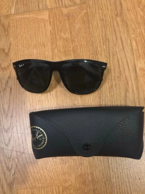 Svarta boyfriends från Ray-Ban - Säljer ett par klassiska svarta solglasögon från Ray-Ban med stilren design. Glasögonen har en snygg och tidlös form, och kommer med ett svart fodral i skinn samt putsduk och instruktionshäfte. Perfekt för dig som vill ha ett ikoniskt accessoar.