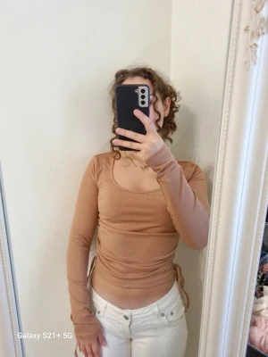 Beige rosa ribbad topp med snörning - Trendig rosa långärmad topp med ribbad struktur och snörning i sidorna för justerbar passform. Toppen har rundad halsringning och är croppad, vilket ger en modern look. Perfekt att styla med både jeans och kjol.