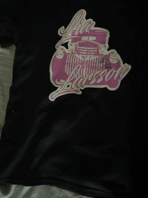 Svart t-shirt med rosa tryck - Snygg svart t-shirt med stort rosa tryck av en bil och texten 'Lia Larsson' på framsidan. Baksidan har turnélista i rosa text. Perfekt för dig som gillar unika bandtröjor och streetstyle.